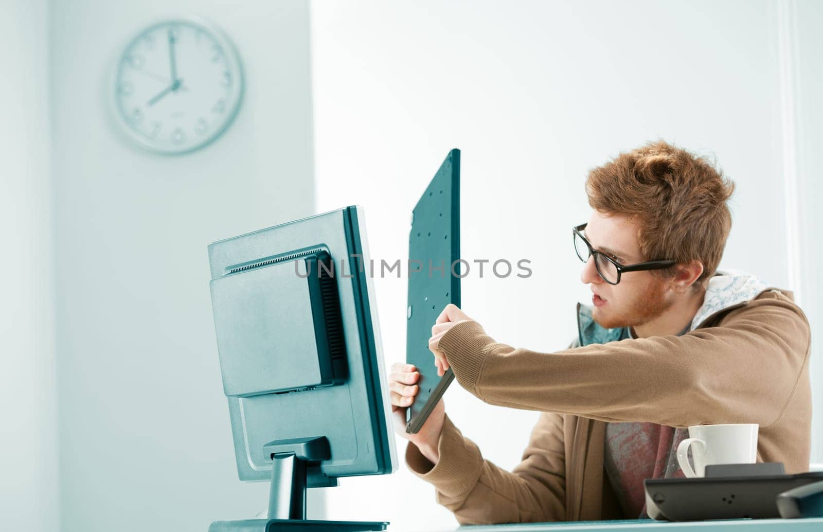 Young red-haired worker angry with his office's computer equipment, tries to destroy the monitor with his keyboard in an extreme gesture of anger and frustration at the unresponsive and uncooperative