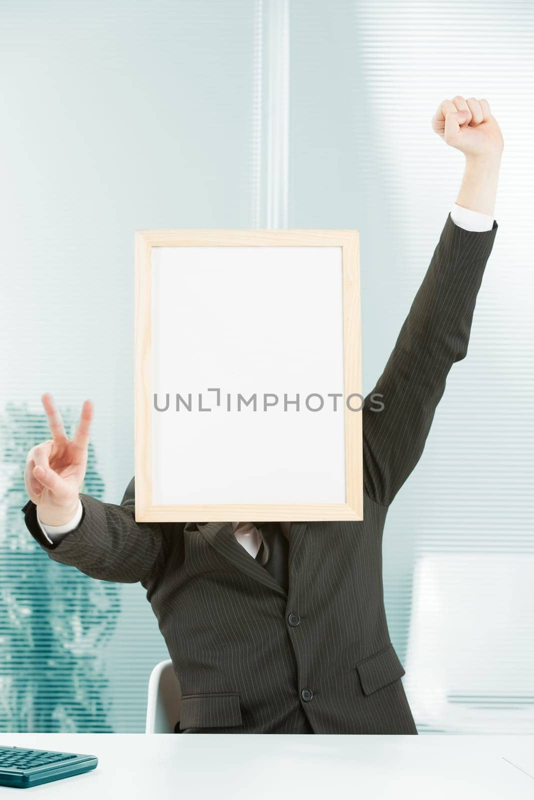 Businessman with sign head has one hand raised in jubilation and with the other forms a victory sign. A computer keyboard can be glimpsed on the desk. There is great enthusiasm at the workplace for th