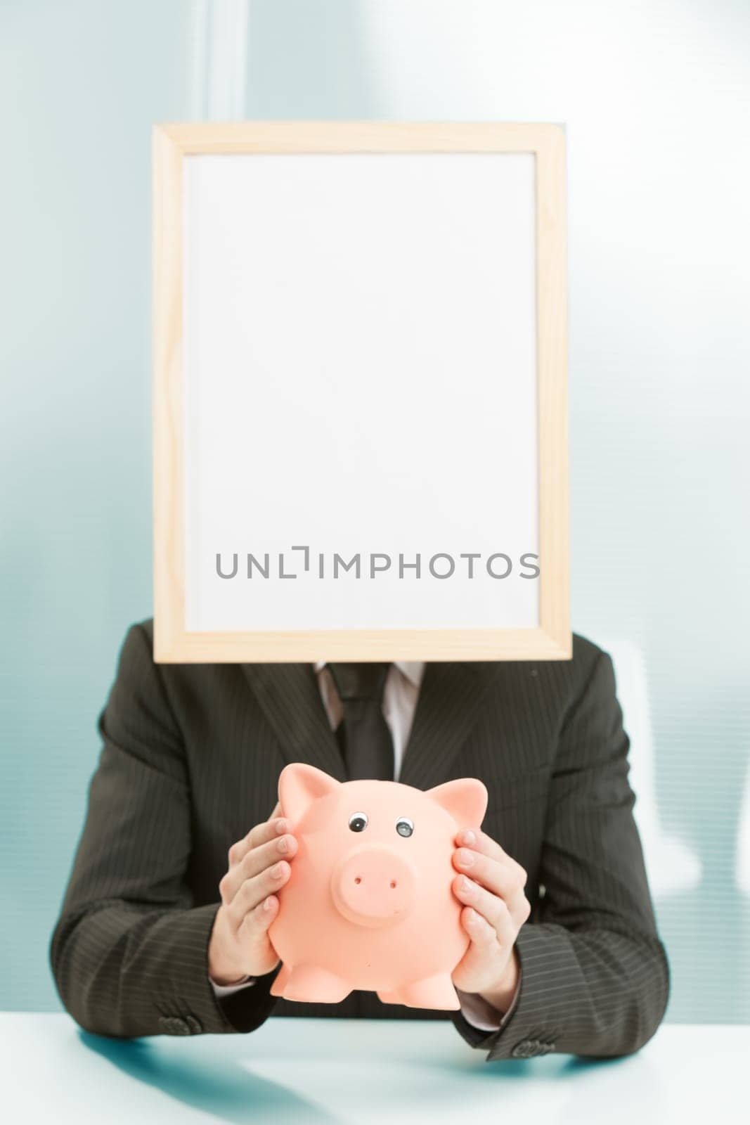 Office workplace, a sign-headed man framed in light wood holds a piggy bank. He holds it carefully, for a concept of savings, spending, investment and generally related to money and business economics