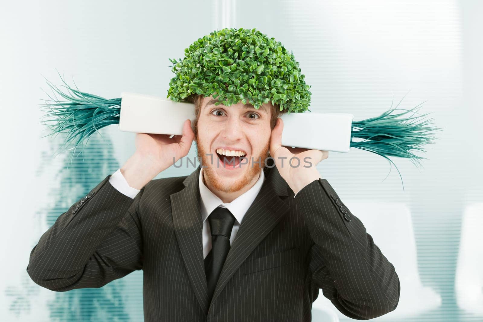 A clerk in an office makes a super smiling and elated face. He has the leafy foliage of a tree instead of hair and pretends to listen to two pots of grass like shells to hear the sound of the sea. Mad