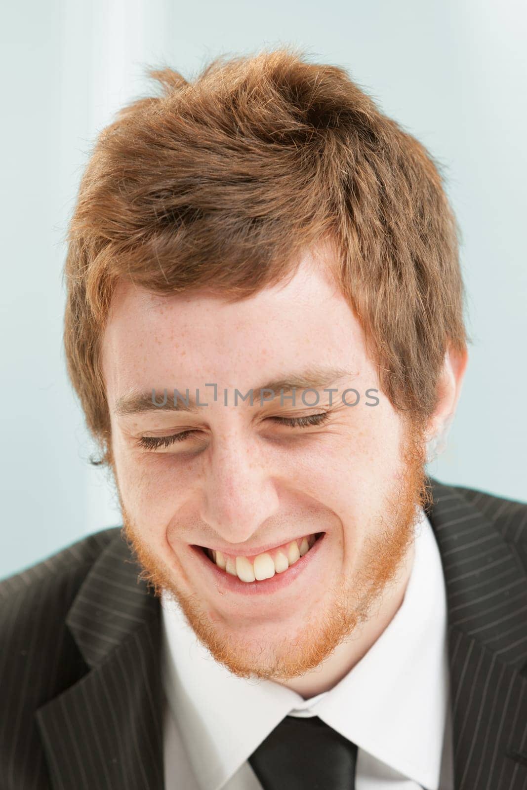 Close-up portrait of young man with short red hair and beard. Positive expression of laughter, his eyes are closed and he is trying not to laugh but he can't help himself and is snickering happily.