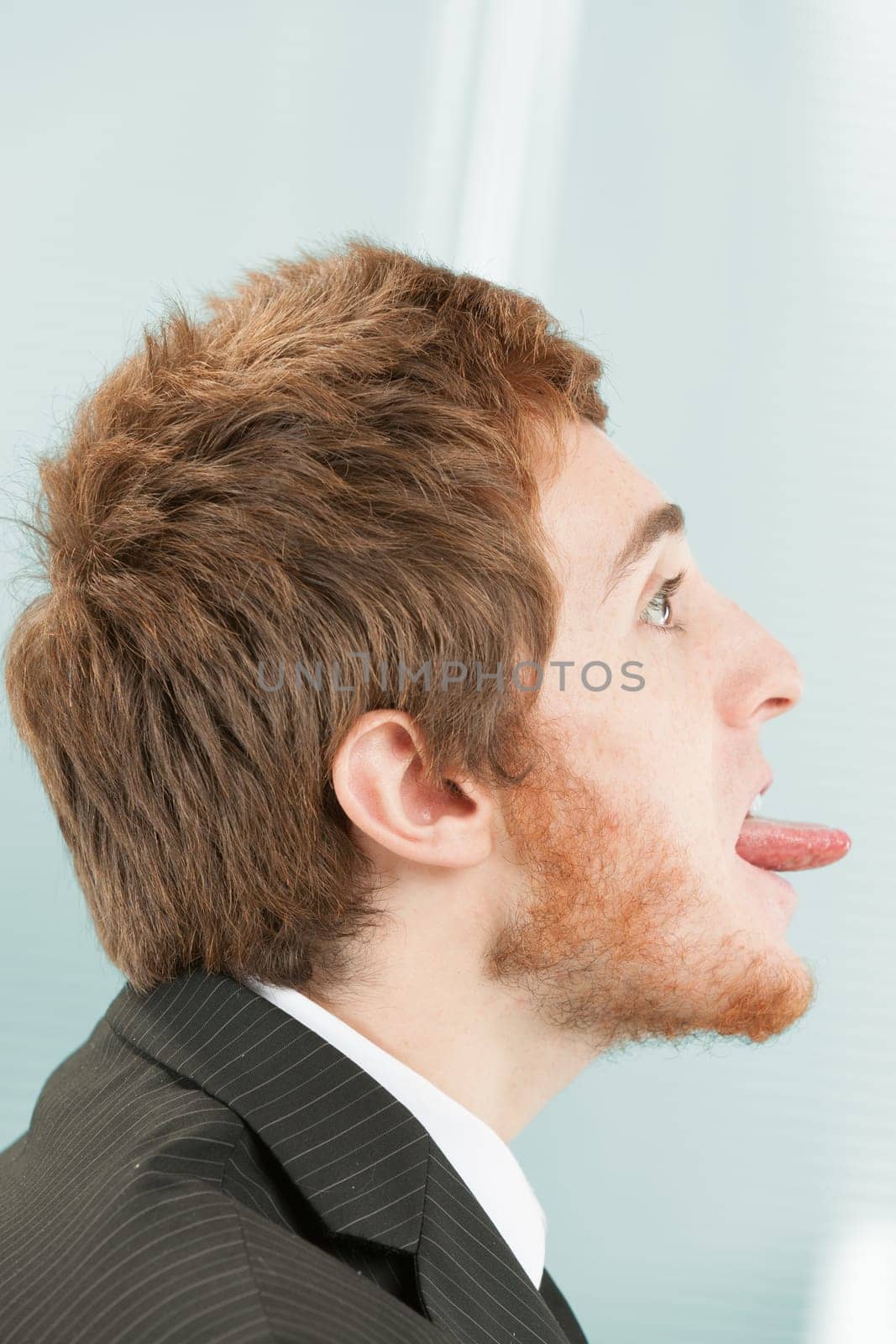 Close-up side portrait of young man with short red hair and beard in suit and tie. Facing to the right (but facing away from himself) he sticks out his entire tongue in defiance, mockery, and sneer at