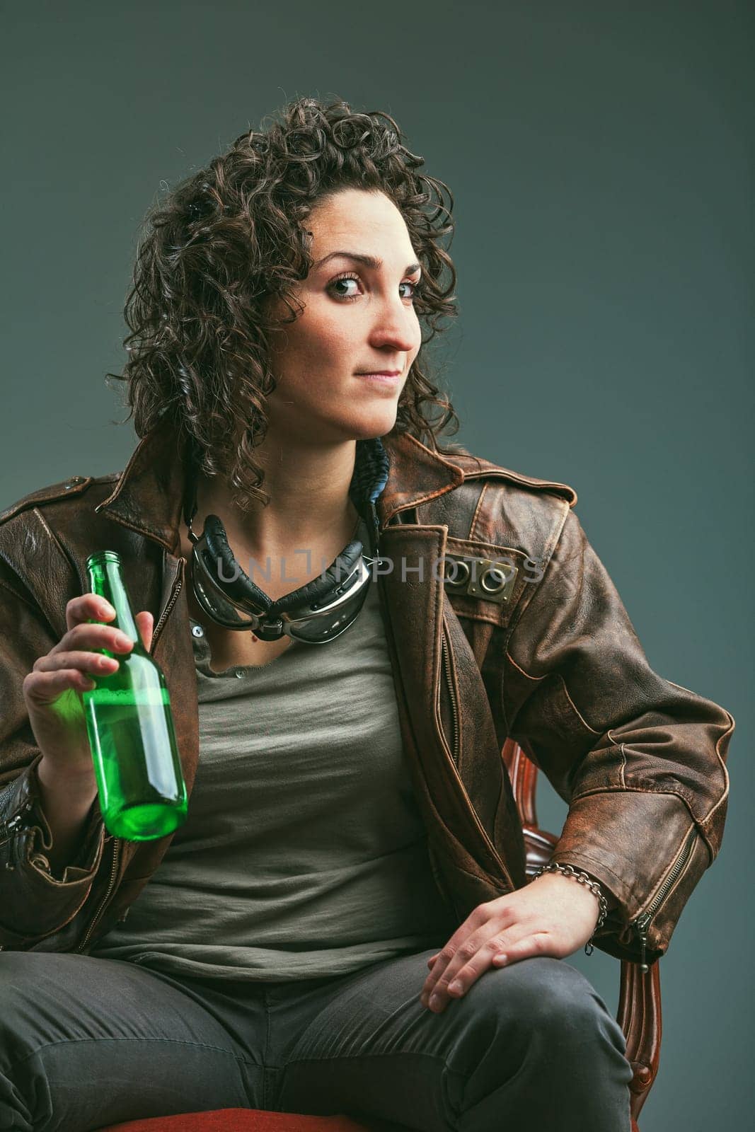 Elegant chair portrait of a biker woman in leather jacket and goggles. She has a defiant look and holds a green glass bottle: enjoying her beer.