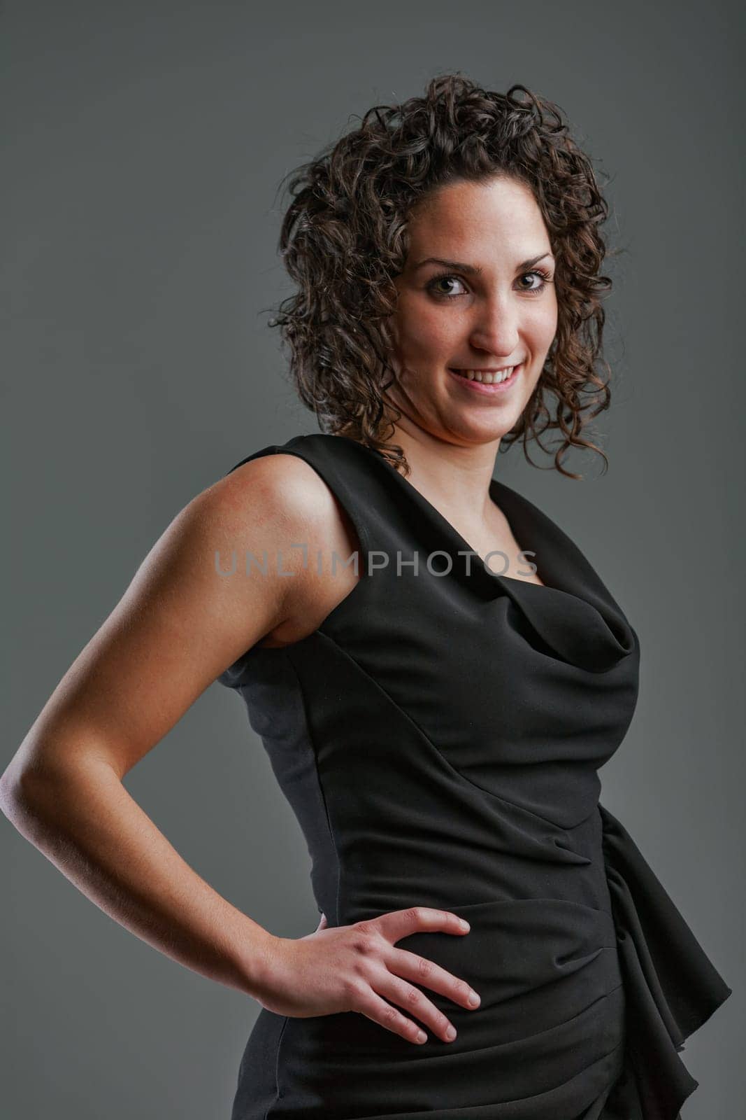 Portrait of a strong woman in a black women's dress with a side bow, she is of sturdy build because women have curves. She has a determined expression and brown curly hair and a hand on her hip.