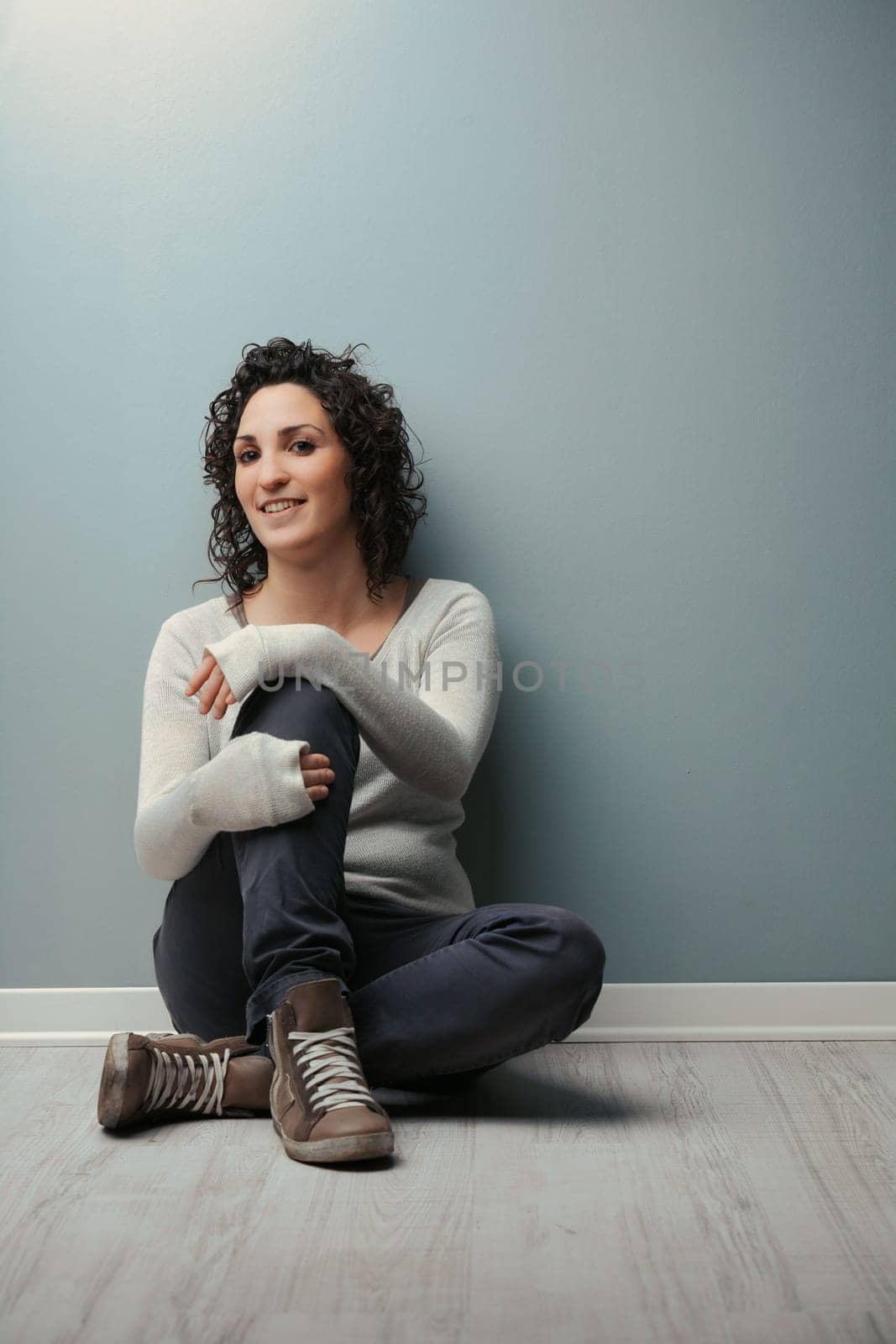 Against her wall, a woman sits with legs crossed, hands on them, in black jeans, shirt, sweater, and sneakers. She's happily serene in her comfy home. A vast COPYSPACE is right above her