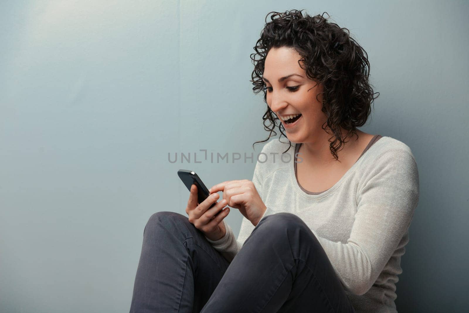 Woman on her home's floor, engrossed with her smartphone's digital content. From videos to social media, every entertainment form is accessible. Donning cozy winter wear like jeans and sweater, she be