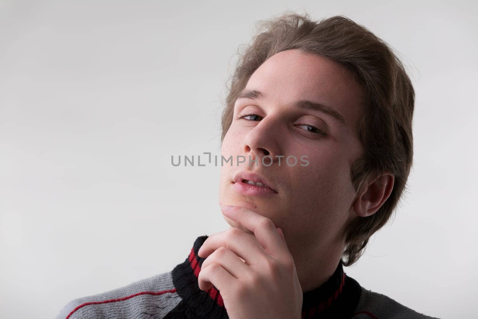 young man with light brown hair, dressed in a gray and black sweater, appears thoughtful with his hand on his chin. As he looks up, it seems he's just had a moment of realization or a brilliant idea