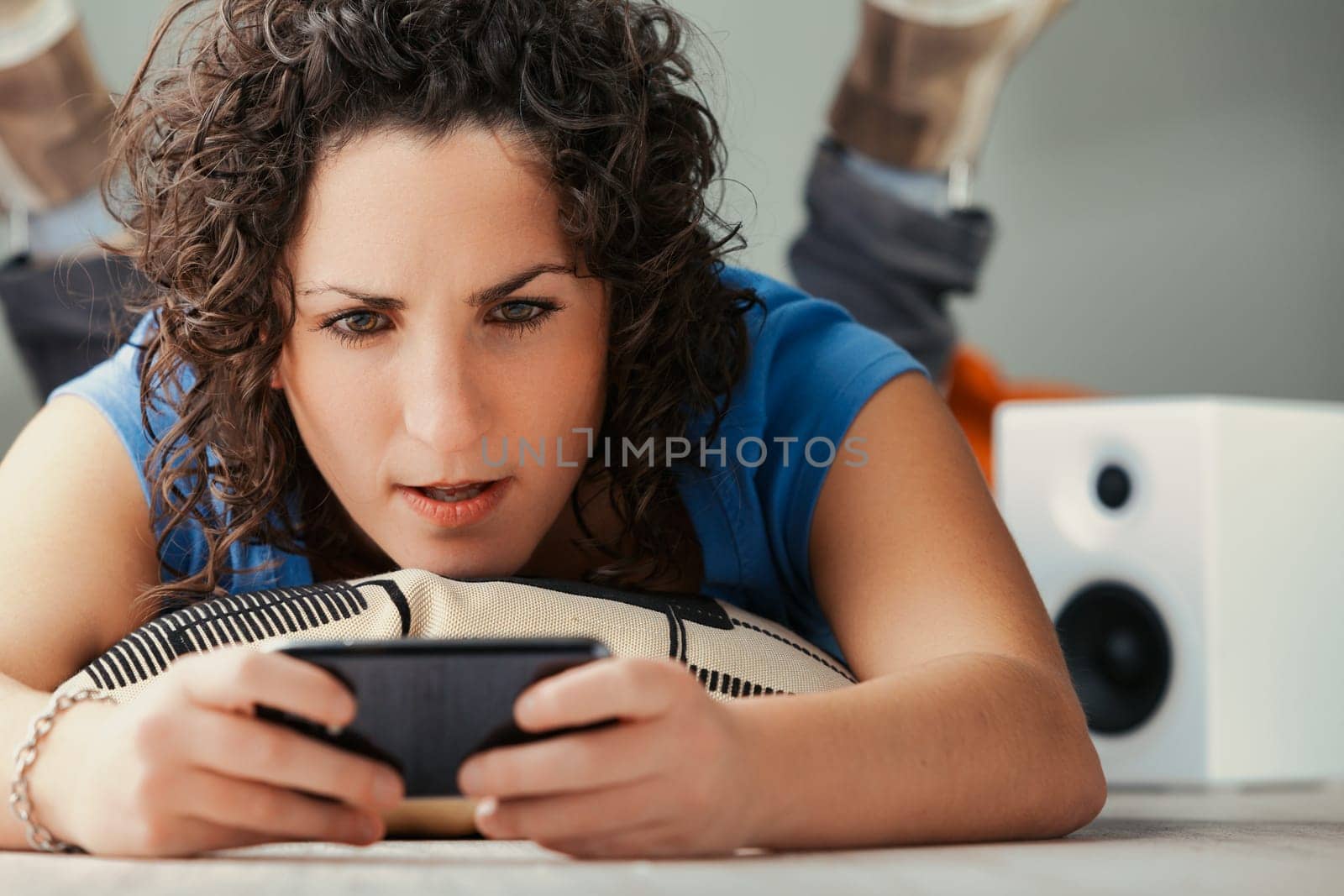 Laid out on cushions, a curly female unwinds at home, engrossed in her phone's universe. Engaging with everything from social networking to adult content, she enjoys her device's multifaceted nature i