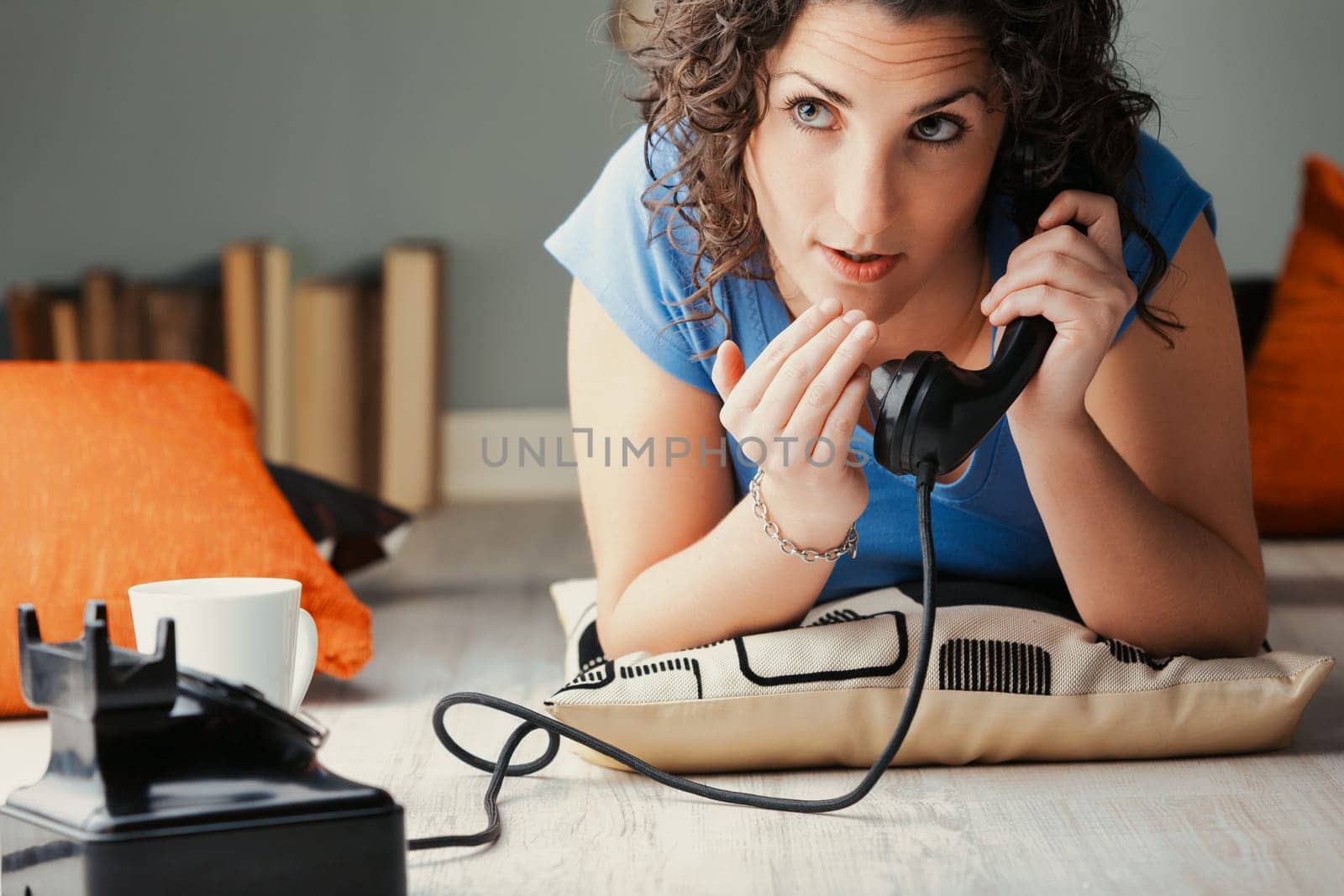 Lounging on the floor amidst cushions, a woman excitedly whispers into a vintage black phone. Amidst the pleasure of gossip, she's oblivious to the fact she'll be discussed next in the rumor mill