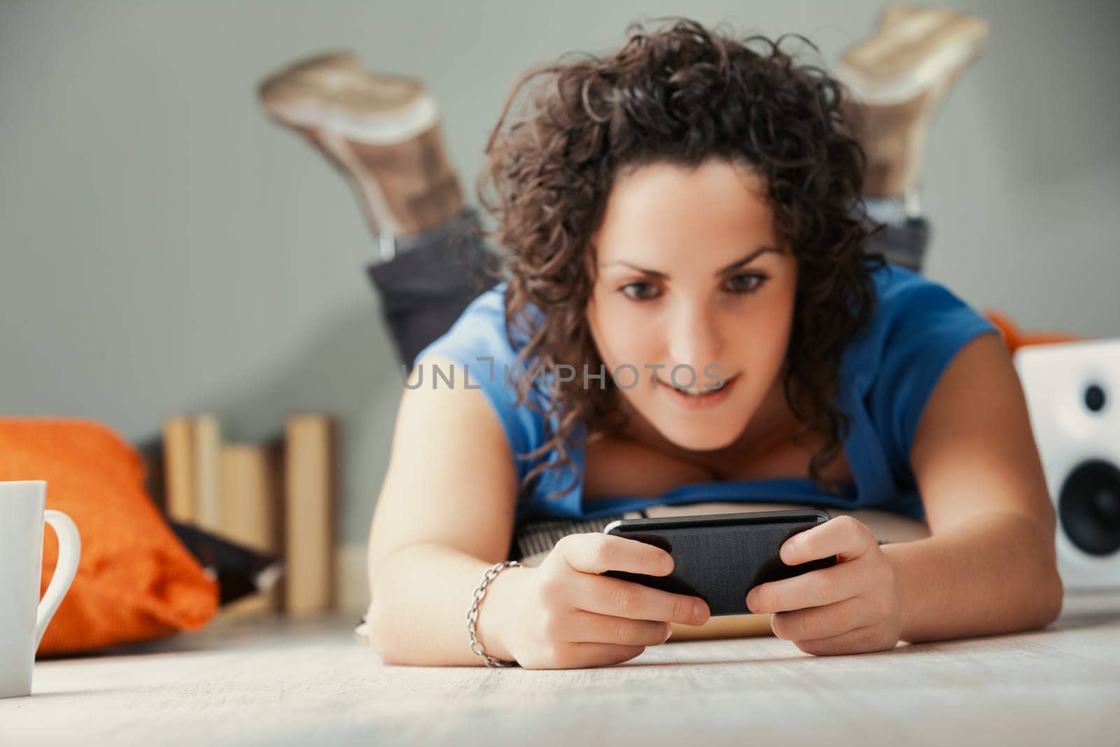 On her cozy home floor, surrounded by cushions, a curly woman immerses herself in her phone's diverse offerings. From social interactions to exploring adult themes, streaming to gaming, she revels in