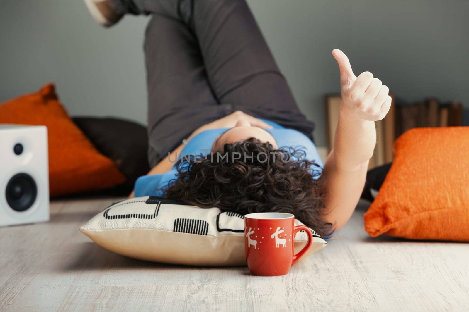 Surrounded by cushions, a young woman with curly hair enjoys her music. Her face is hidden, but her outstretched thumb signals her contentment. Beside her, a holiday mug adds a touch of festivity