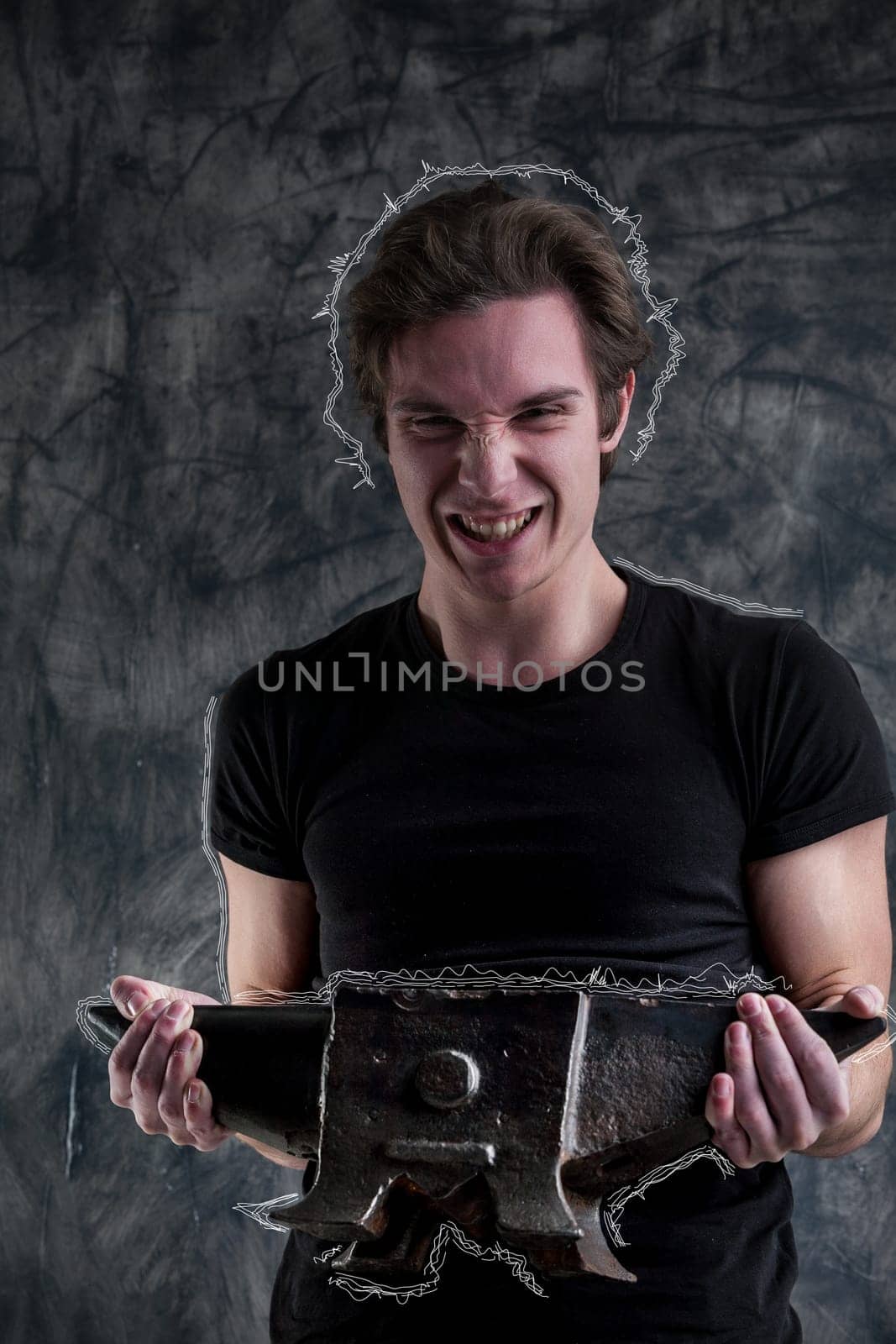 On a dramatic grunge-black backdrop, a young man in a black shirt lifts an anvil with notable effort, yet succeeds. His robust muscles aid him. Even strained, joy emerges through his laughter