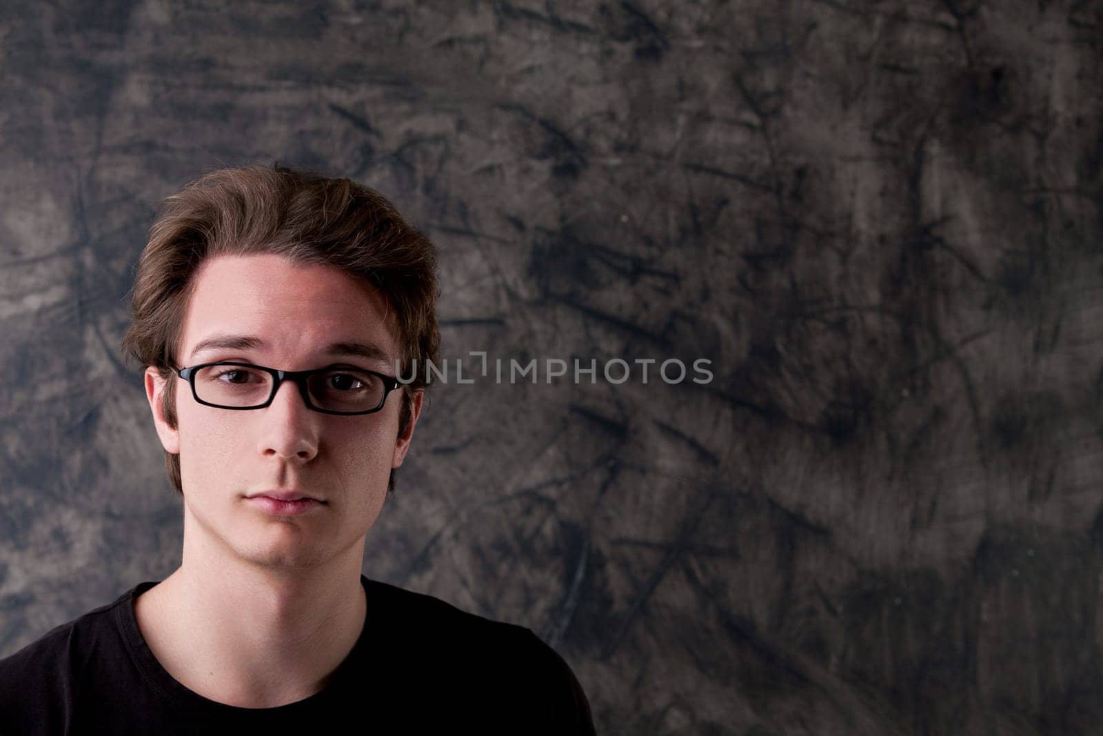 Young man with glasses in front of a wiped chalkboard. Ample copyspace available. His nerdy expression conveys skepticism towards imprecise or illogical remarks, urging for more informed insights