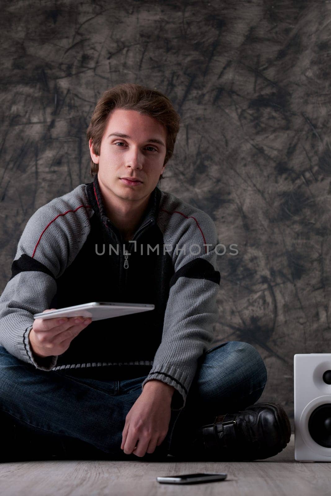 Young man in a grey and black sweater stands indoors, near white hi-fi speakers and a smartphone. He extends a digital tablet, promoting mobile tech, apps, and online tools for work and entertainment