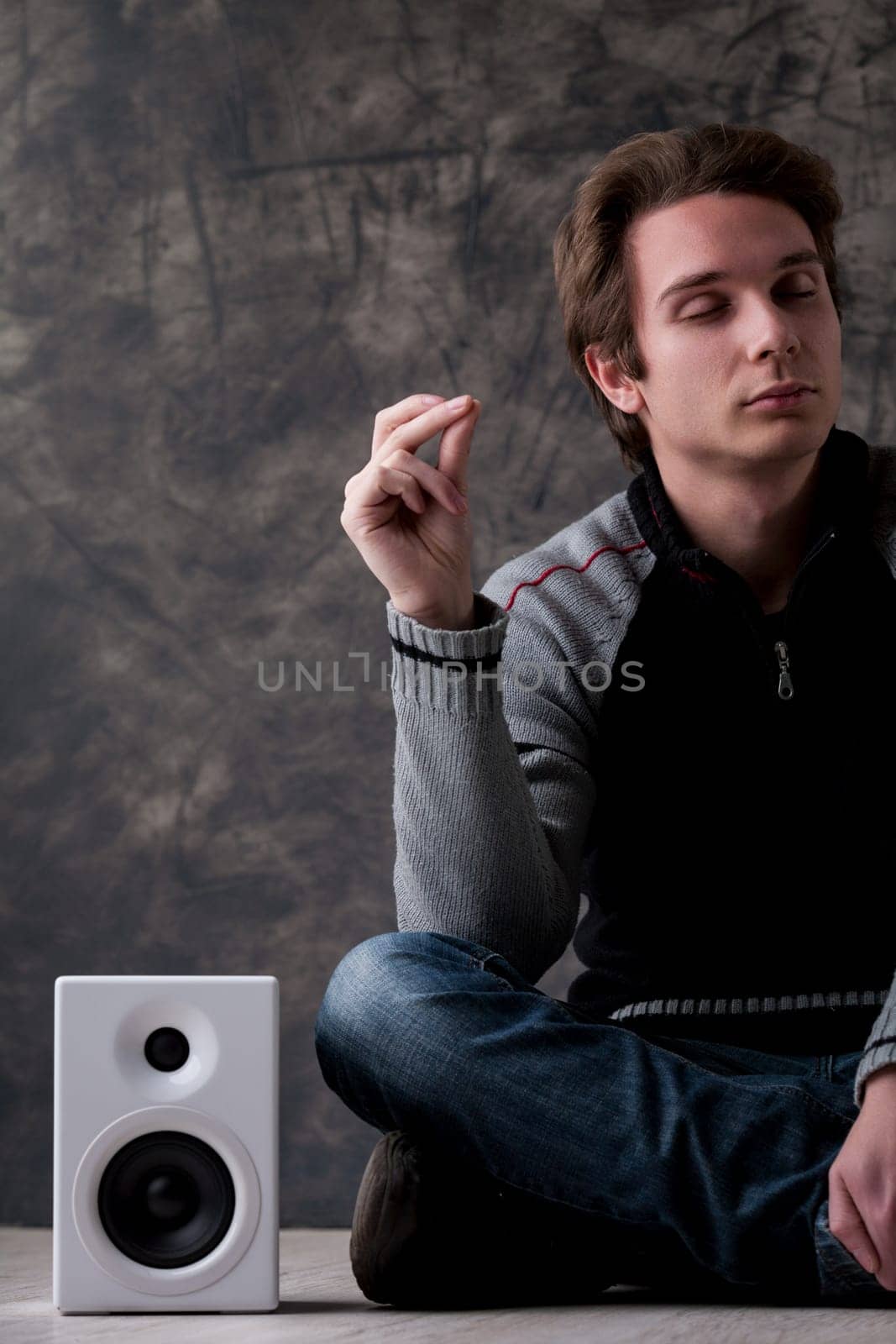 Young man in a black and grey sweater sits cross-legged near white hi-fi speakers, snapping fingers to the rhythm with eyes closed. He enjoys the groove, unaware it's machine-generated upsampled music