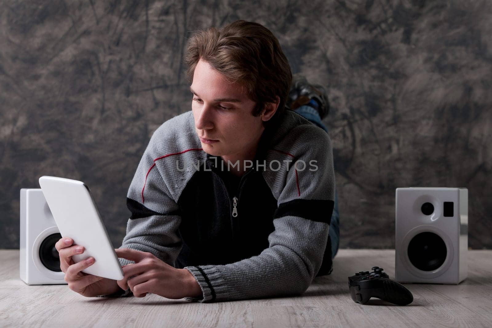 Young man lies on the floor with a white digital tablet, gaming controller, and speakers nearby. Digital entertainment is great if you don't have time to go out. Content is vast, but ownership is fadi