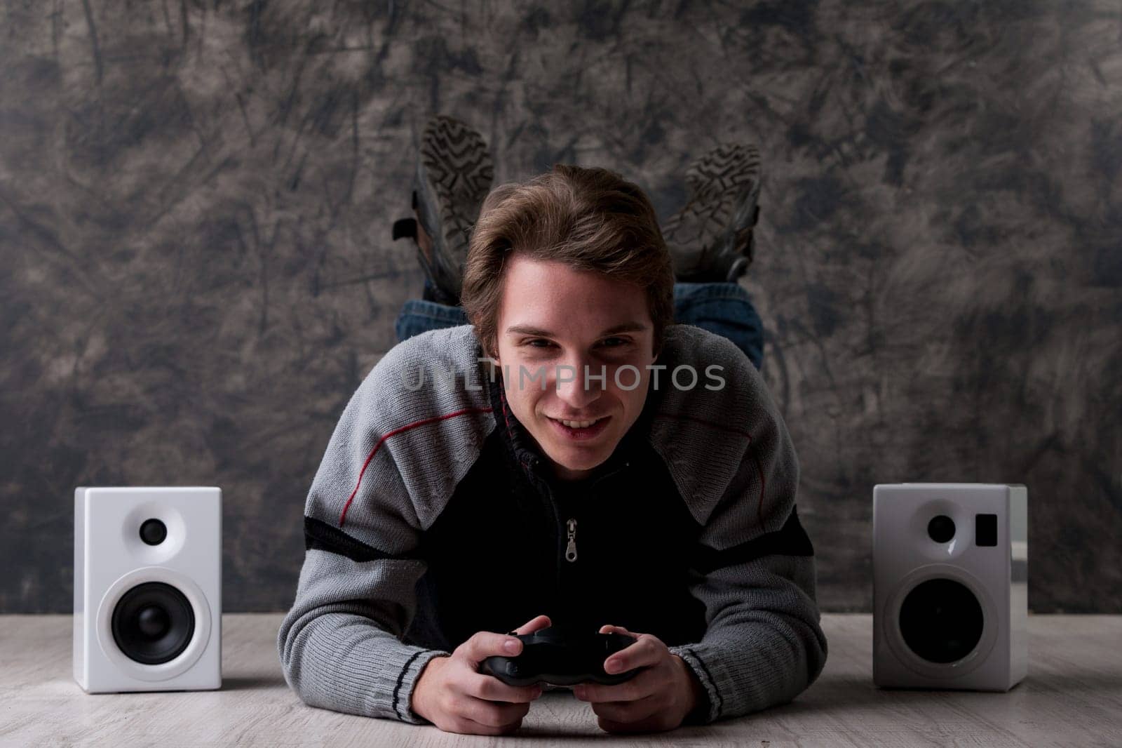 Young man lies on the floor with a video game controller and high-quality stereo speakers. Eager to win every online tournament and become the best in e-sports, he's keen, fierce, and excited. 'Eat my