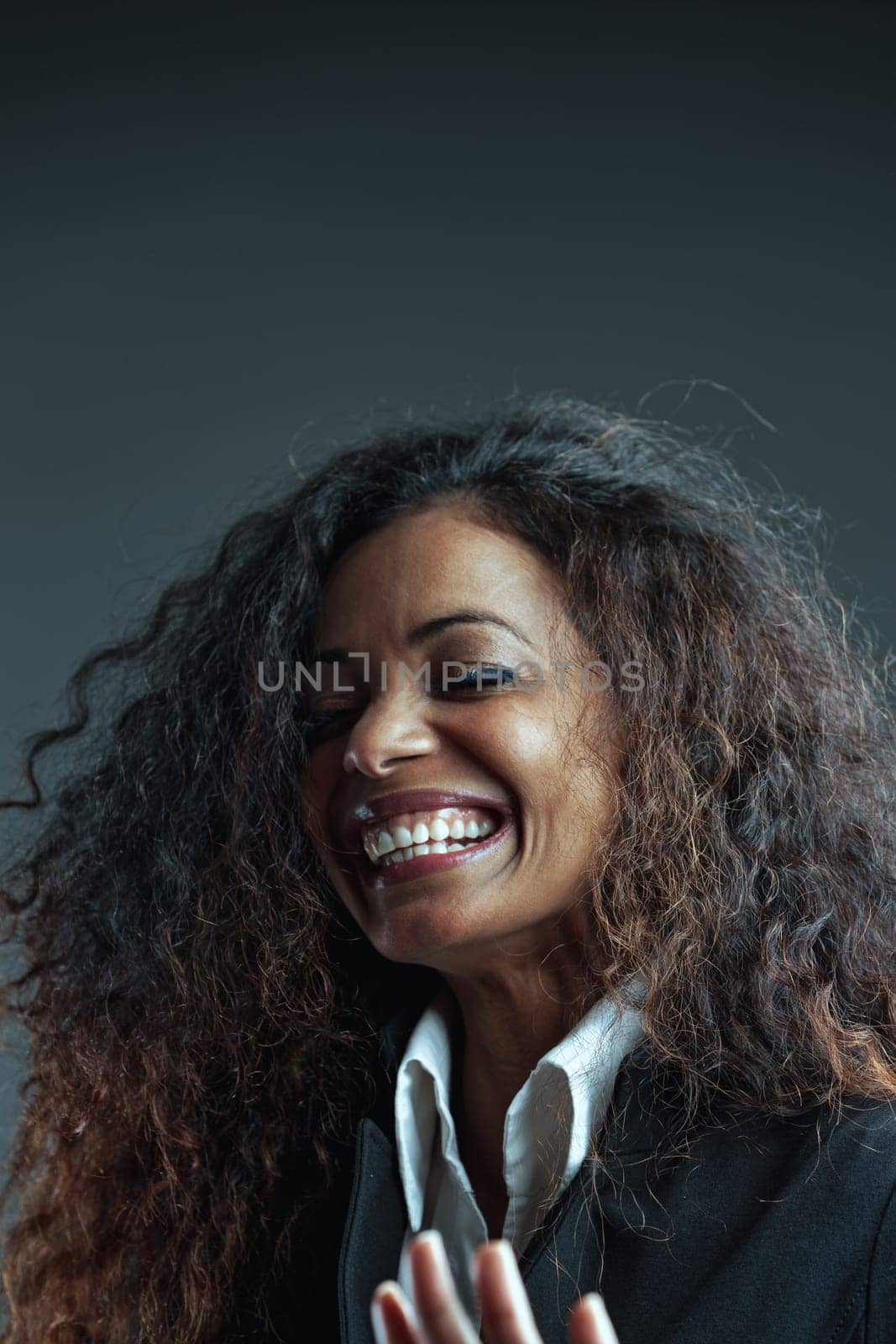 Portrait of Black-skinned Woman with long wavy hair, in elegant white shirt and black jacket, caught in the midst of laughter or an intense moment of joy. Perfect teeth, eyes closed.