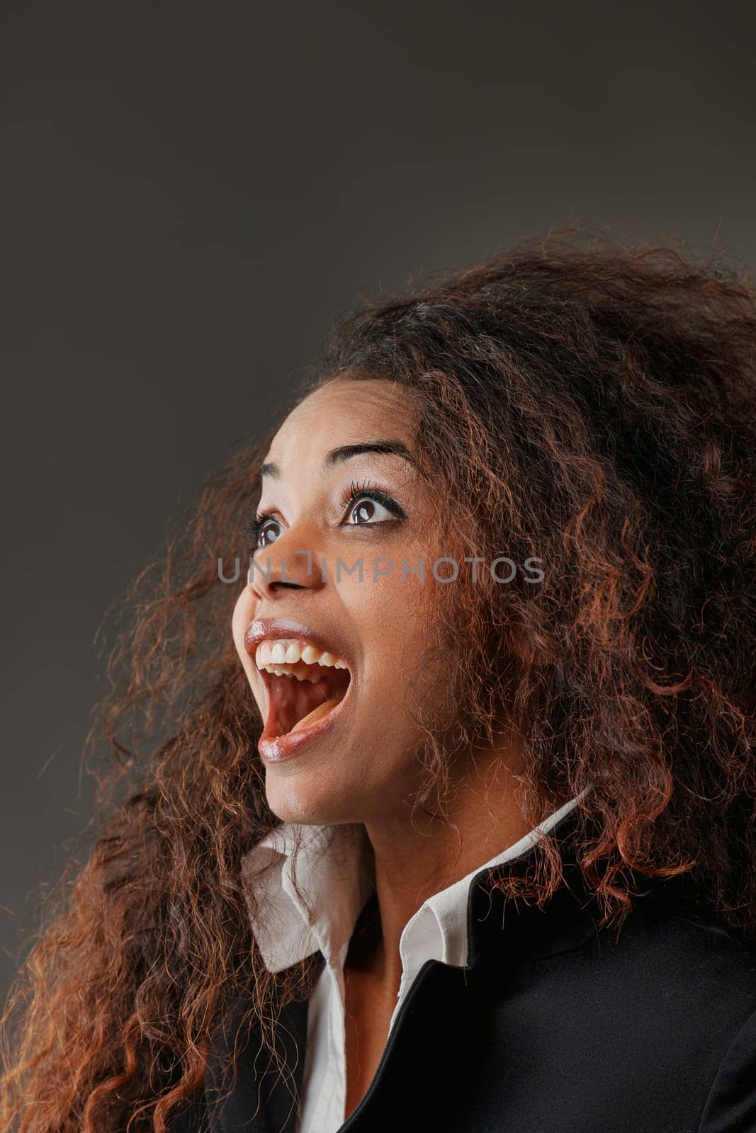 Exuding joy, a stunning black woman with luscious lips gazes upwards, astonished by something positively amazing. Wearing a white shirt, black jacket, and boasting long curly hair, her joy is infectio