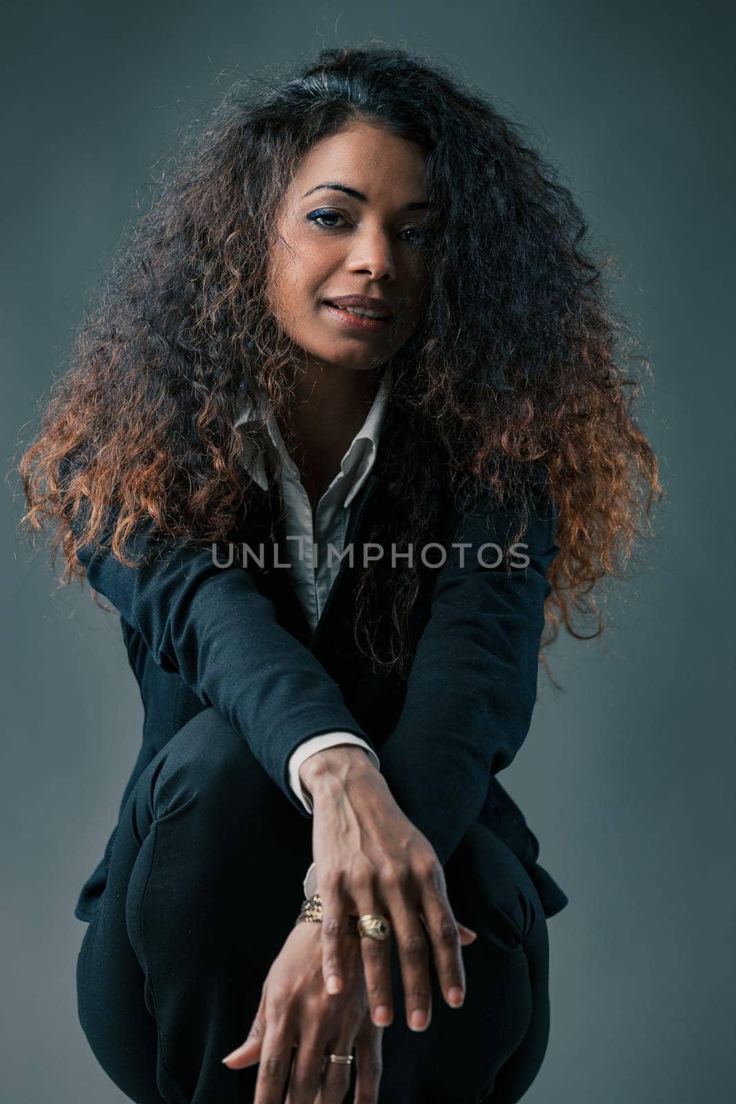 Reflecting on trials, woman in black stands out. Curly mane, striking features define her. Fatigue hints in her smile strength undeterred. Against a somber setting
