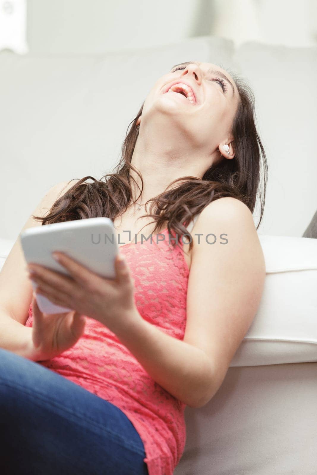 A woman on the couch in her living room with a digital tablet in hand. She's wearing a red tank top and jeans, and she looks happy and intelligent. Her house is bright, and she's either checking her a
