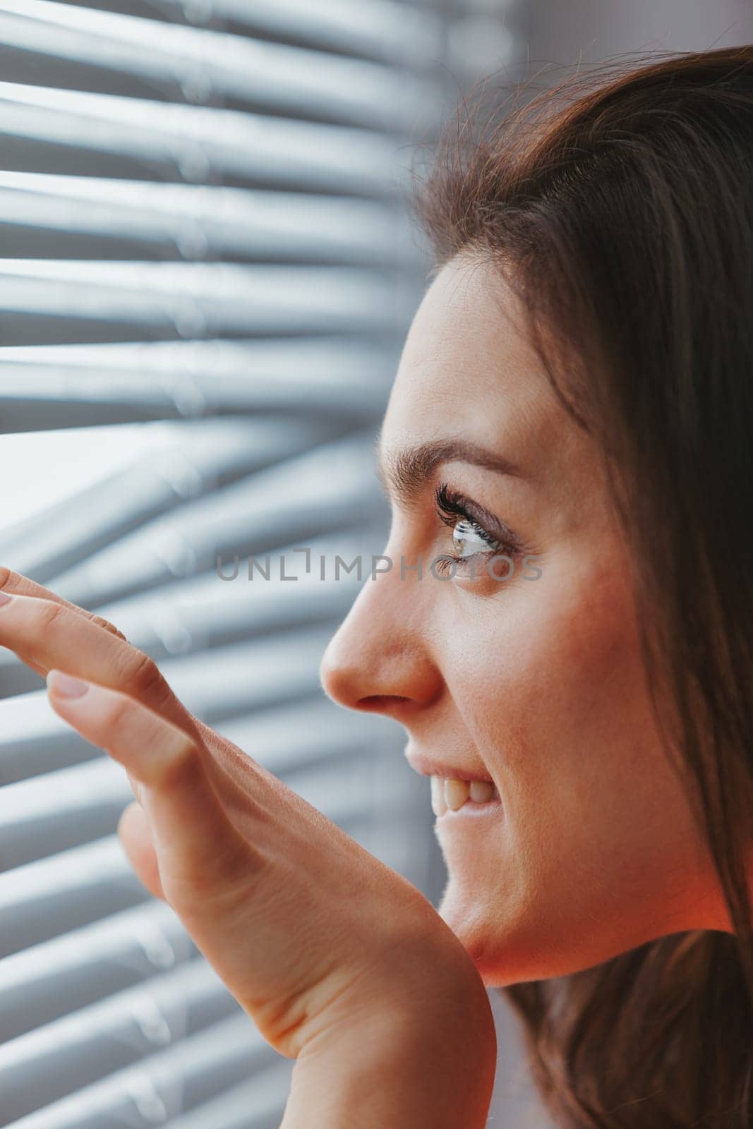 A close-up profile portrait of a young woman who is smiling but cautious as she peers through the slats of the blinds to see what's outside. Is it a beautiful day? Is it daytime or nighttime? Am I saf