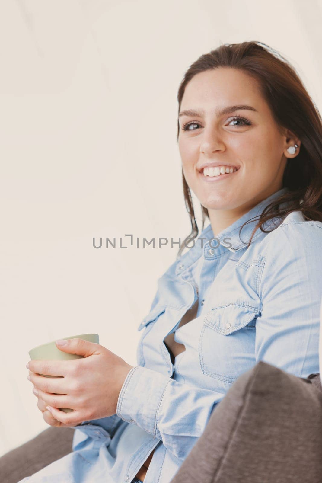 A happy and smiling woman sits on her white couch at home, enjoying her free time with style. She sips a hot drink from a green mug while wearing a blue denim shirt and skirt. Sunlight streams in thro