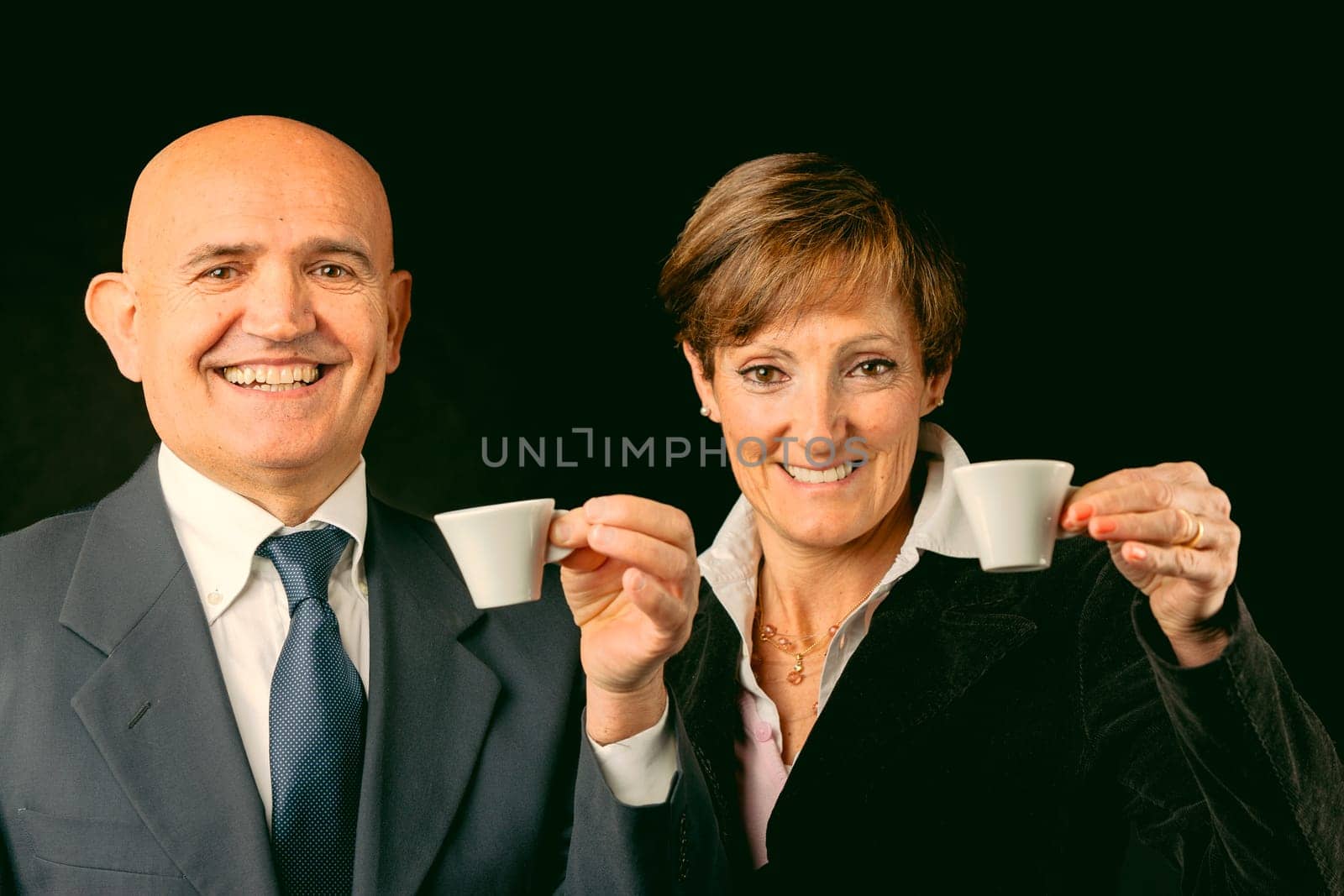 Portrayed elegantly against black, two wealthy elderly enjoy Italian espresso on their European voyage. While he's bald with jacket and shirt, she flaunts boyish red hair. Both toast to the health of