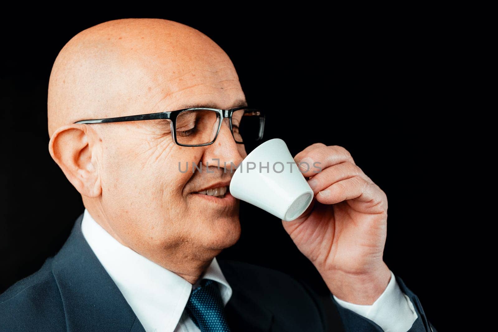aged bald man is captured up-close, relishing genuine Italian espresso from a suitably-sized cup. Dressed in a dark jacket and white shirt, with black glasses on, he's a picture of contentment. Note: