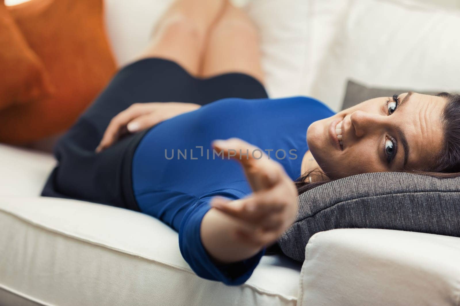A woman is lounging on her white sofa in her free time, surrounded by orange pillows. She is wearing a blue shirt and a black miniskirt. She extends her hand towards someone she loves, inviting them t