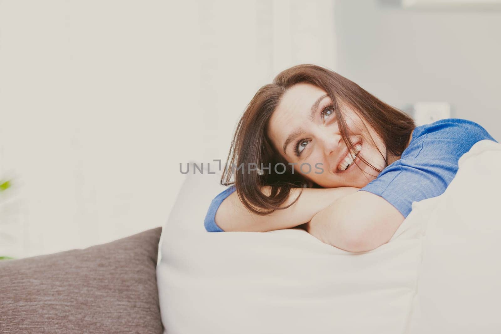 A young, happy, and dreamy woman smiles with her head resting on her crossed arms, which are in turn leaning against the cushion of the sofa. She imagines, invents, and reasons. She imagines a way to