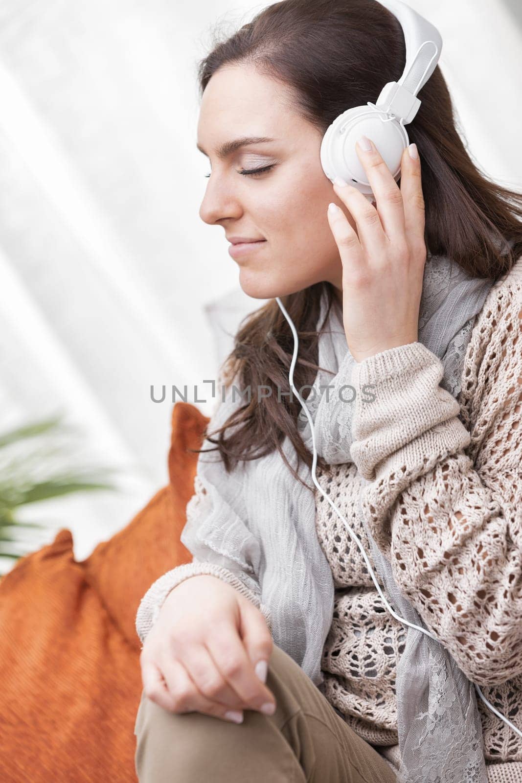 A woman dressed in shades of brown is sitting barefoot on her home sofa. She's listening to music with white headphones, or maybe to the radio or an interesting podcast. Relaxing and entertaining at h