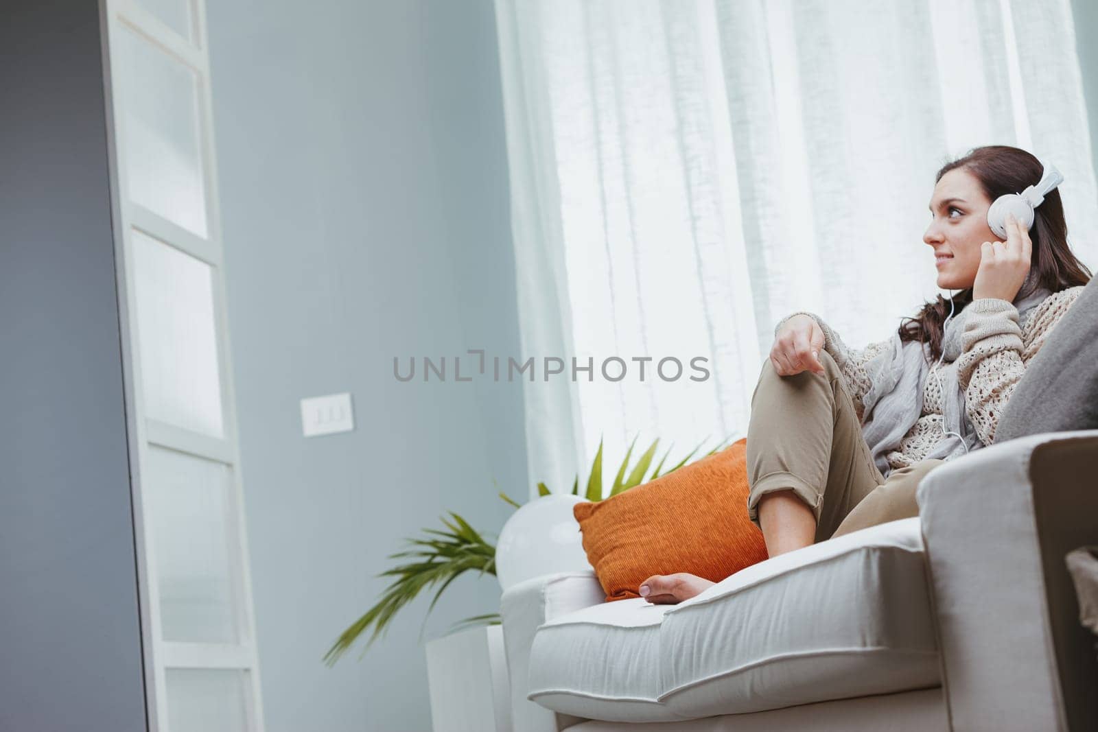 A woman dressed in shades of brown is sitting barefoot on her home sofa. She's listening to music with white headphones, or maybe to the radio or an interesting podcast. Relaxing and entertaining at h