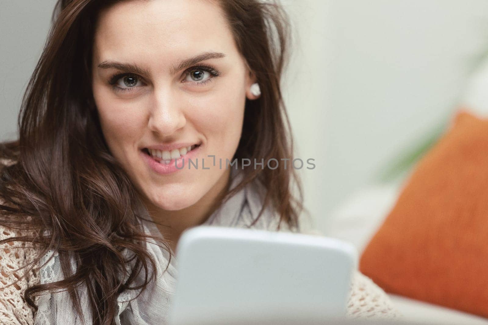Portrait of a young woman with beautiful eyes that convey intelligence, holding a white digital tablet. She sits on the carpet at the foot of the sofa, enjoying culture and entertainment from the inte