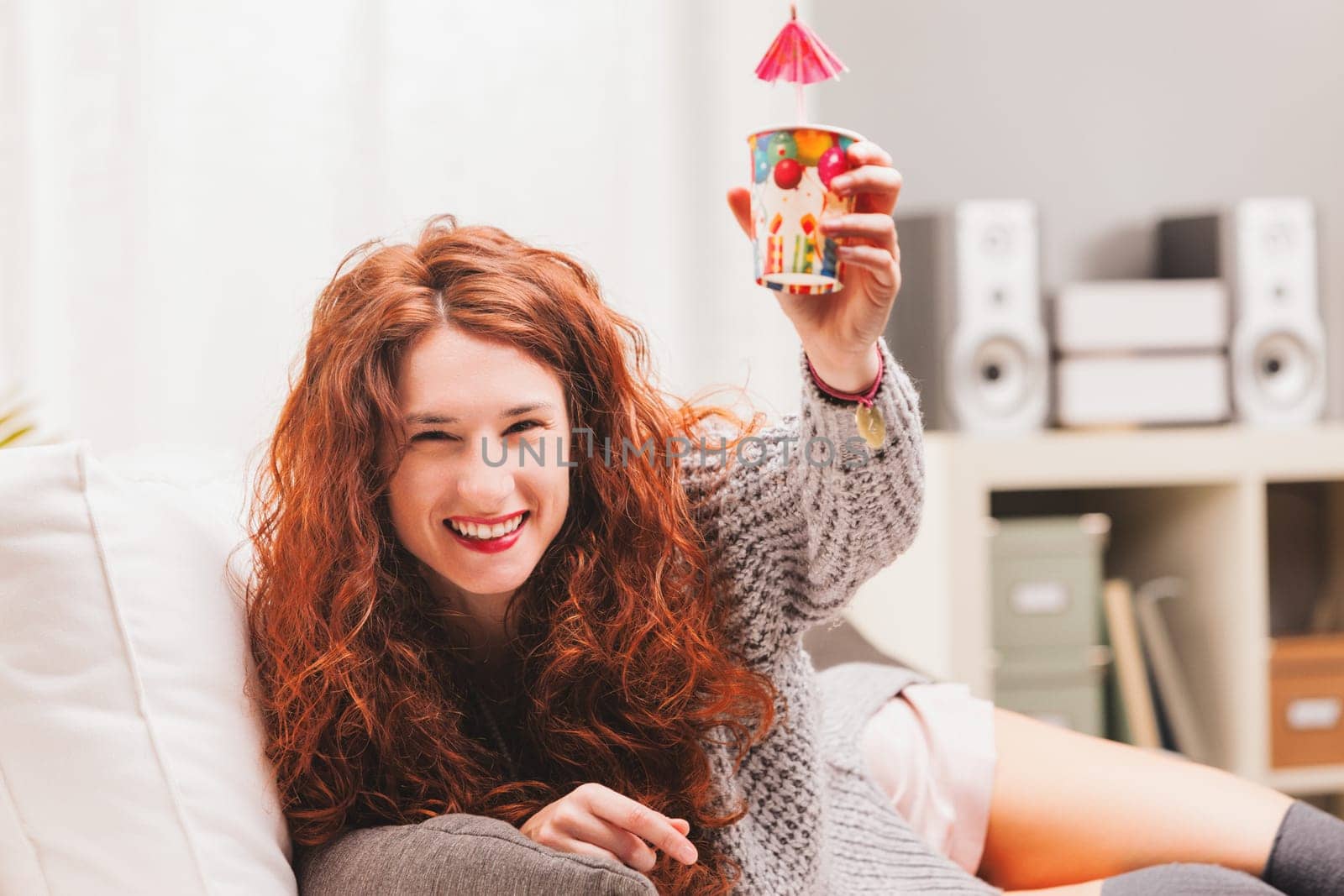 Embodying a carefree lifestyle, a radiant redhead on a white sofa revels in festivities. In a mix of intimate apparel and a woolen top, she beams with joy, raising celebratory items in cheer