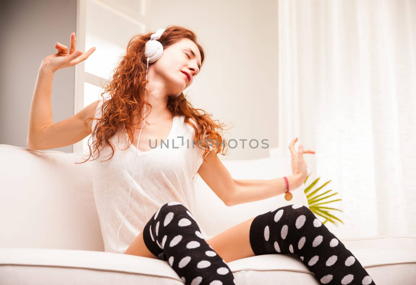 Embracing her love for music, a woman with fiery long hair dances, sings, and feels its essence. Relaxed in her attire, she cherishes her time on a white sofa at home
