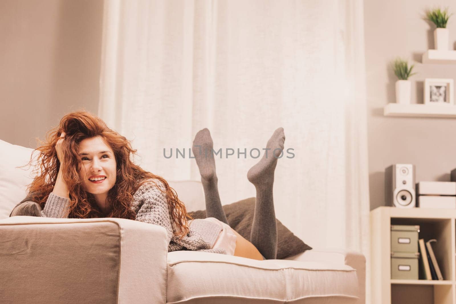 Dreaming of weekends or vacations, possible dates or career progress, a woman lays relaxed in her intimate wear, sweater, and knee-high stockings on her couch. Her joy and satisfaction are evident, co