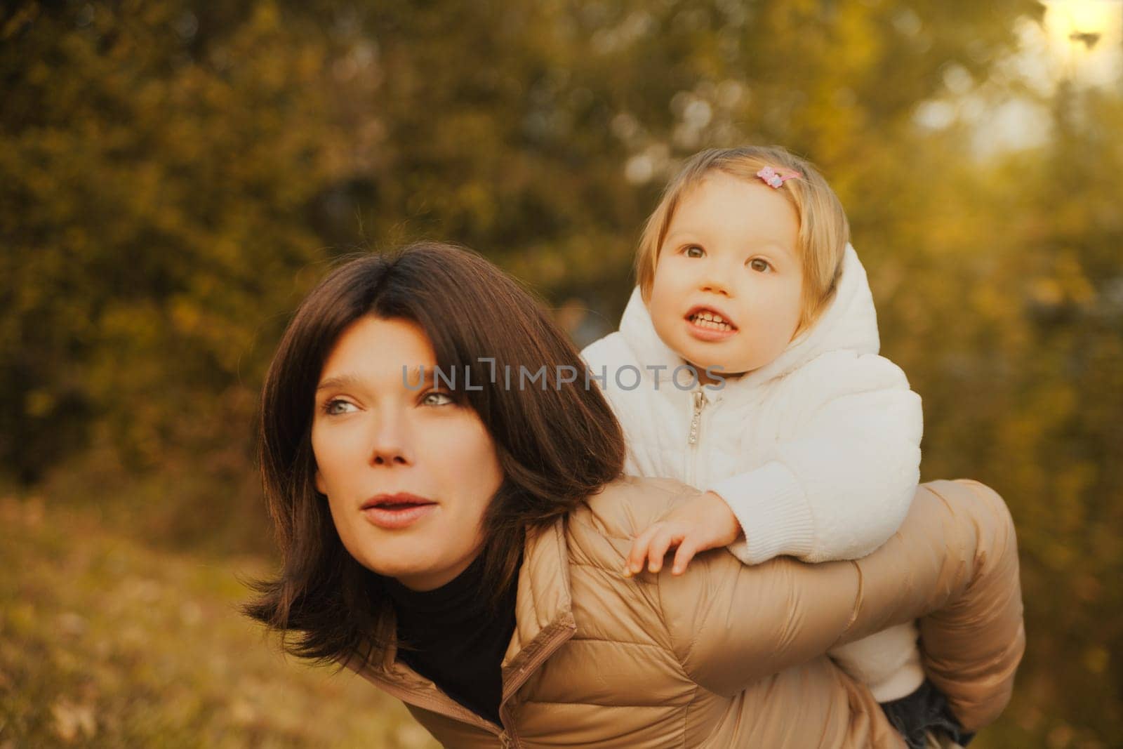 Enjoying the outdoors, a blonde child climbs atop her lovely mother's shoulders. They're both snug against autumn's chill, sharing a day of boundless love and company