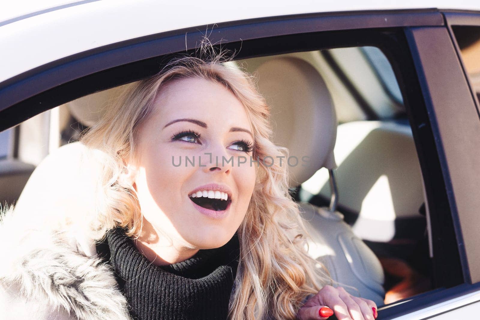 In a quaint European city, an elegant blonde woman with flowing curls drives her stylish car. Her makeup and manicure flawless, she smiles and poses, highlighting her bewitching eyes, sun-shaded at ti