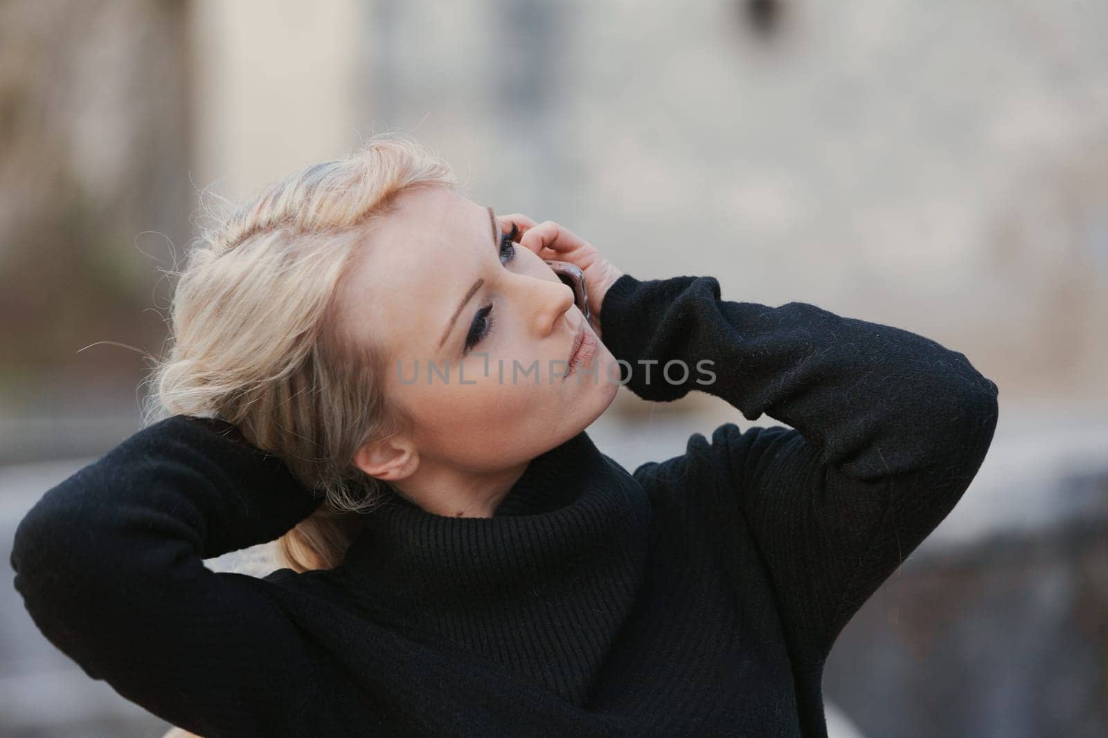 Young woman with long, curly, perfectly permed blonde hair, perfect makeup and manicure, is a tourist in a European city with medieval villages. Using her phone to talk, chat, or access Wi-Fi for hist