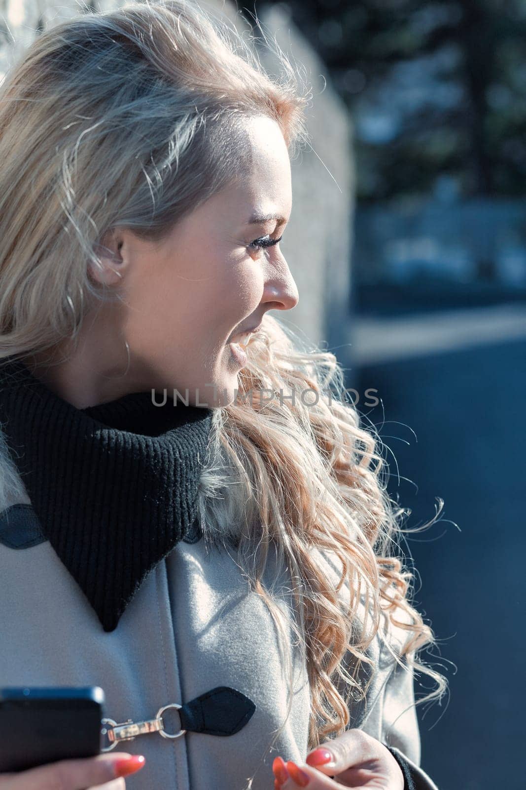 Young woman with long, curly, perfectly permed blonde hair, perfect makeup and manicure, is a tourist in a European city with medieval villages. Using her phone to talk, chat, or access Wi-Fi for hist