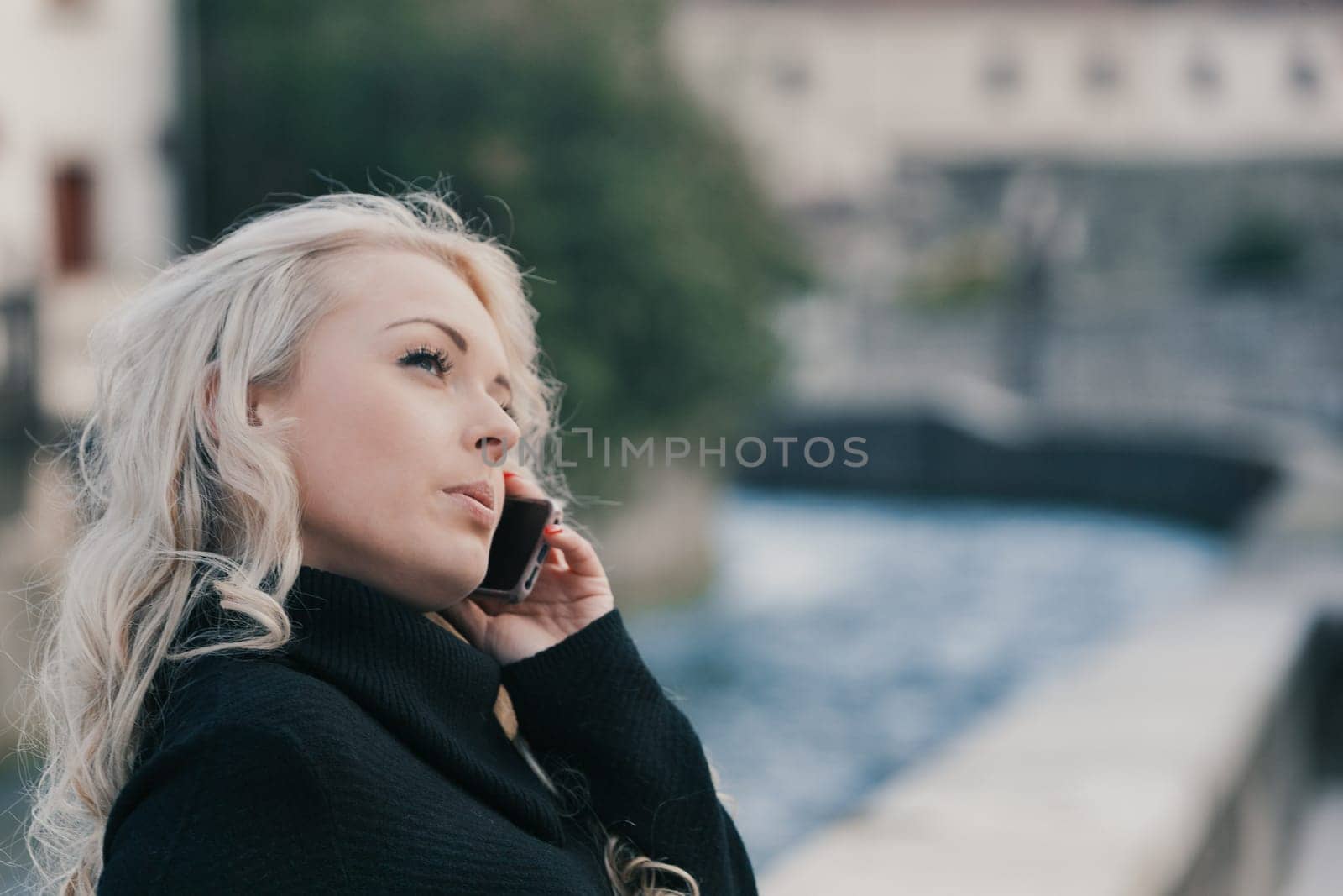 Young woman with long, curly, perfectly permed blonde hair, perfect makeup and manicure, is a tourist in a European city with medieval villages. Using her phone to talk, chat, or access Wi-Fi for hist