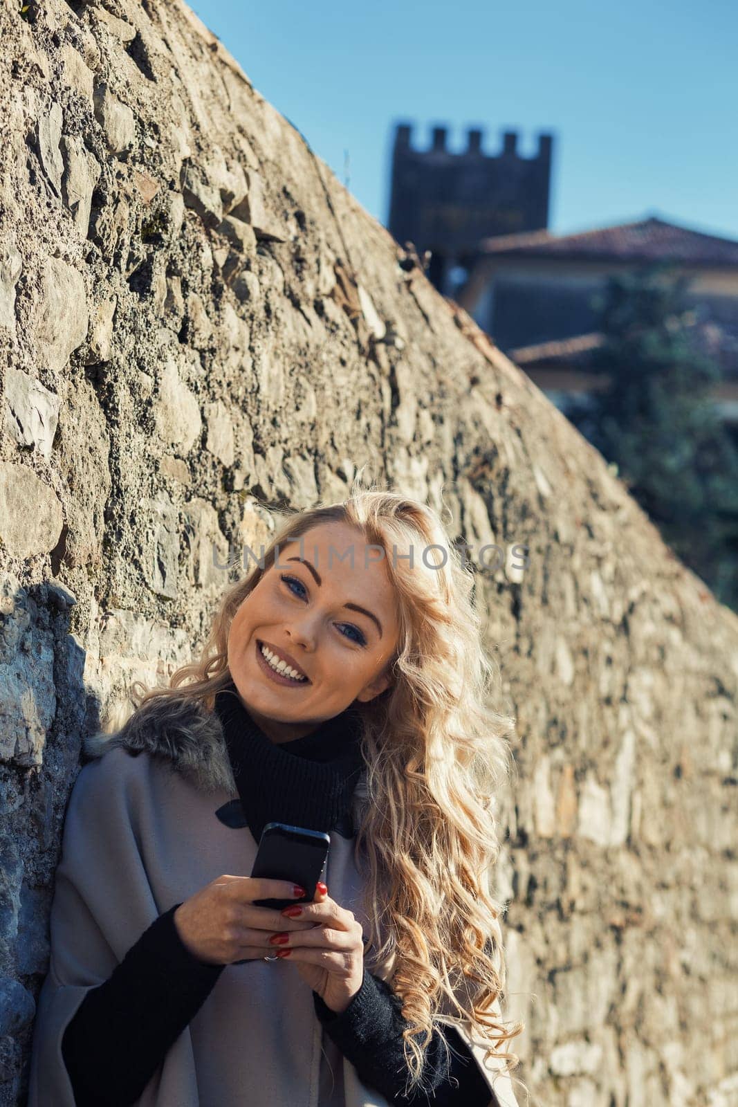 Young woman with long, curly, perfectly permed blonde hair, perfect makeup and manicure, is a tourist in a European city with medieval villages. Using her phone to talk, chat, or access Wi-Fi for hist