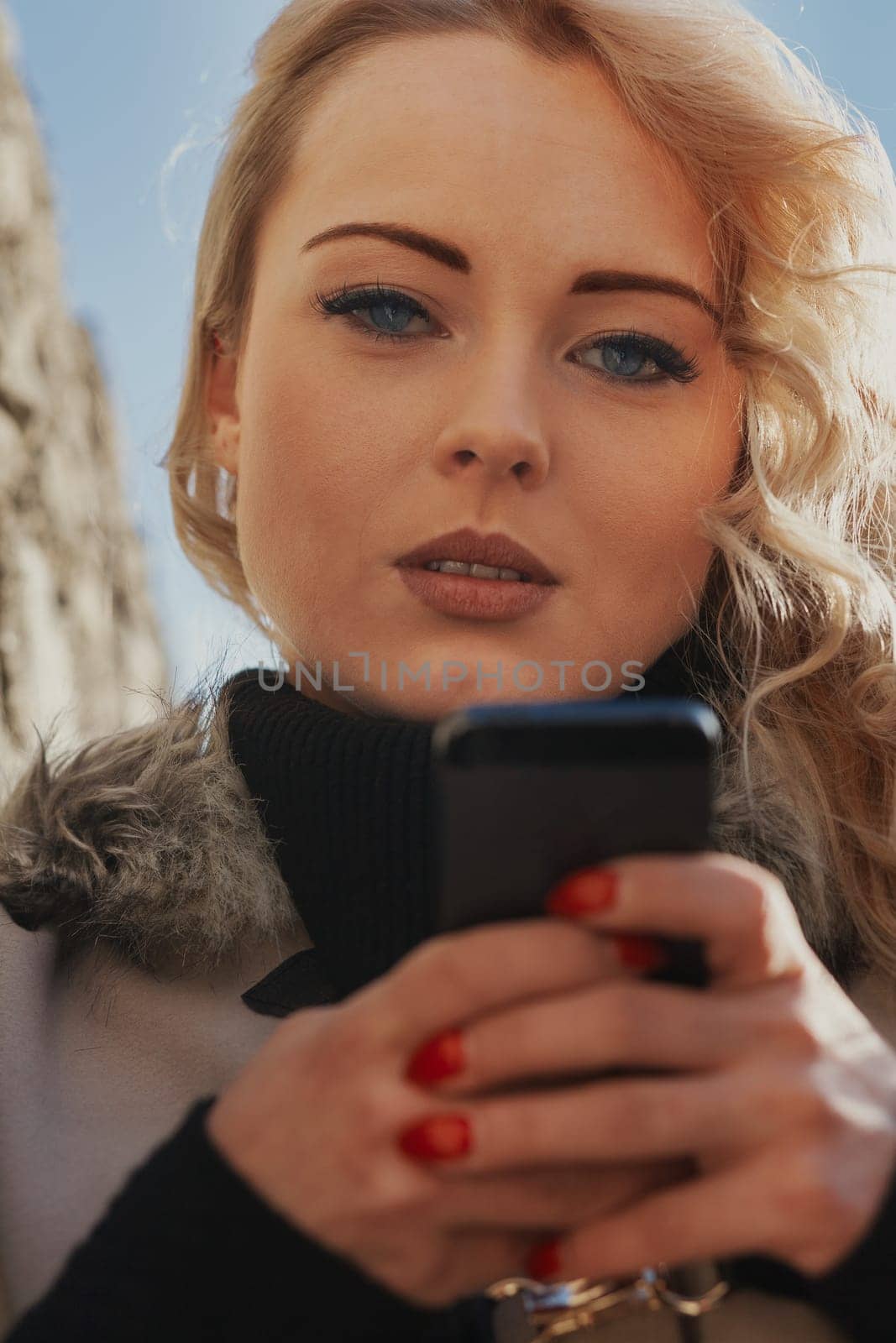Young woman with long, curly, perfectly permed blonde hair, perfect makeup and manicure, is a tourist in a European city with medieval villages. Using her phone to talk, chat, or access Wi-Fi for hist