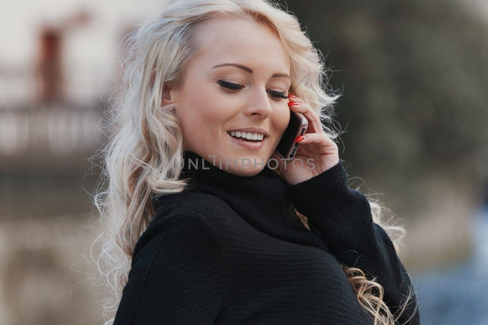 Young woman with long, curly, perfectly permed blonde hair, perfect makeup and manicure, is a tourist in a European city with medieval villages. Using her phone to talk, chat, or access Wi-Fi for hist