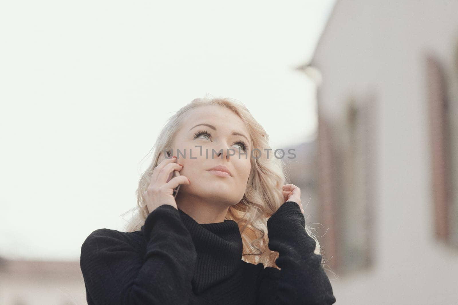 Young woman with long, curly, perfectly permed blonde hair, perfect makeup and manicure, is a tourist in a European city with medieval villages. Using her phone to talk, chat, or access Wi-Fi for hist