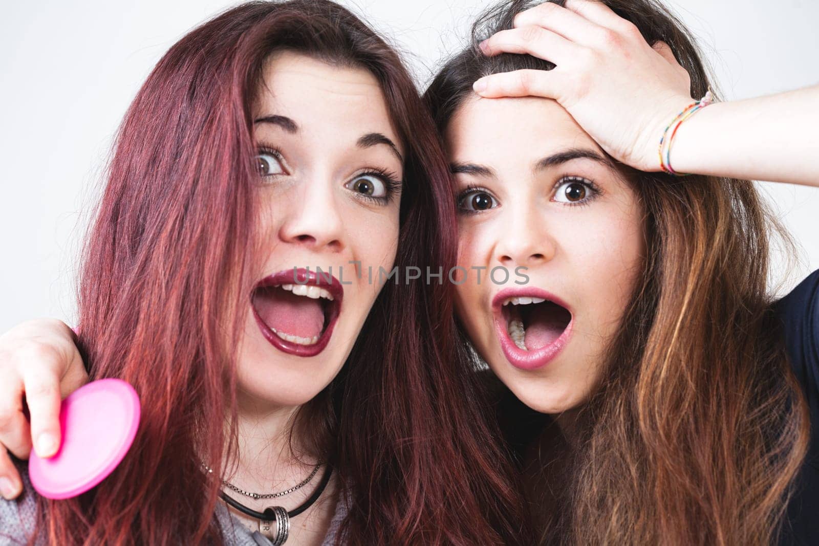 Two youthful friends, impeccably made-up and with flowing hair, let loose, making funny faces. They revel in the carefree moments and their playful expressions shine through