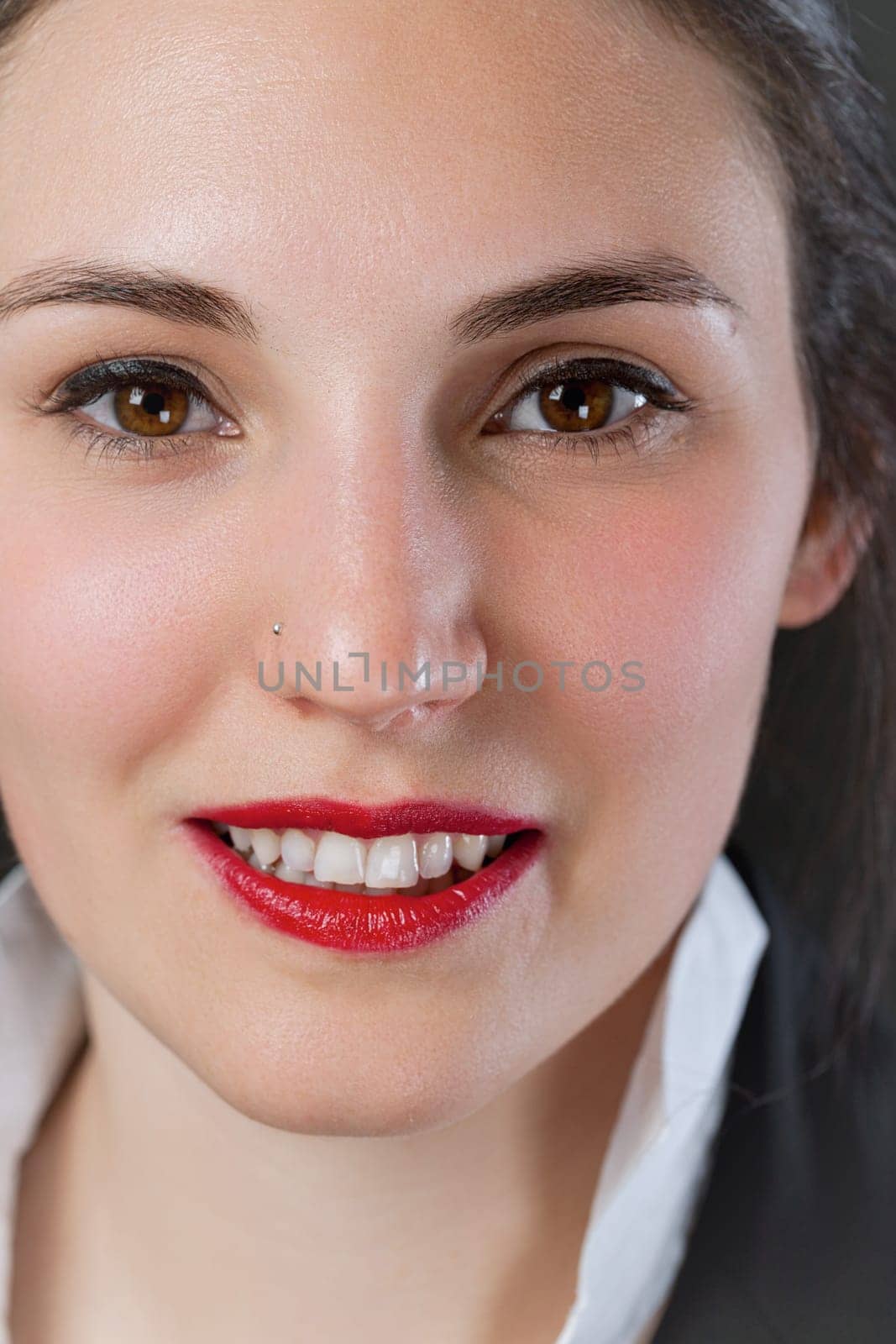 Frontal close-up portrait of a woman, red lipstick and visible white collar beneath a black jacket. Shes sensual and sweet, but annoy her and you'll find out her real strength. Be very careful