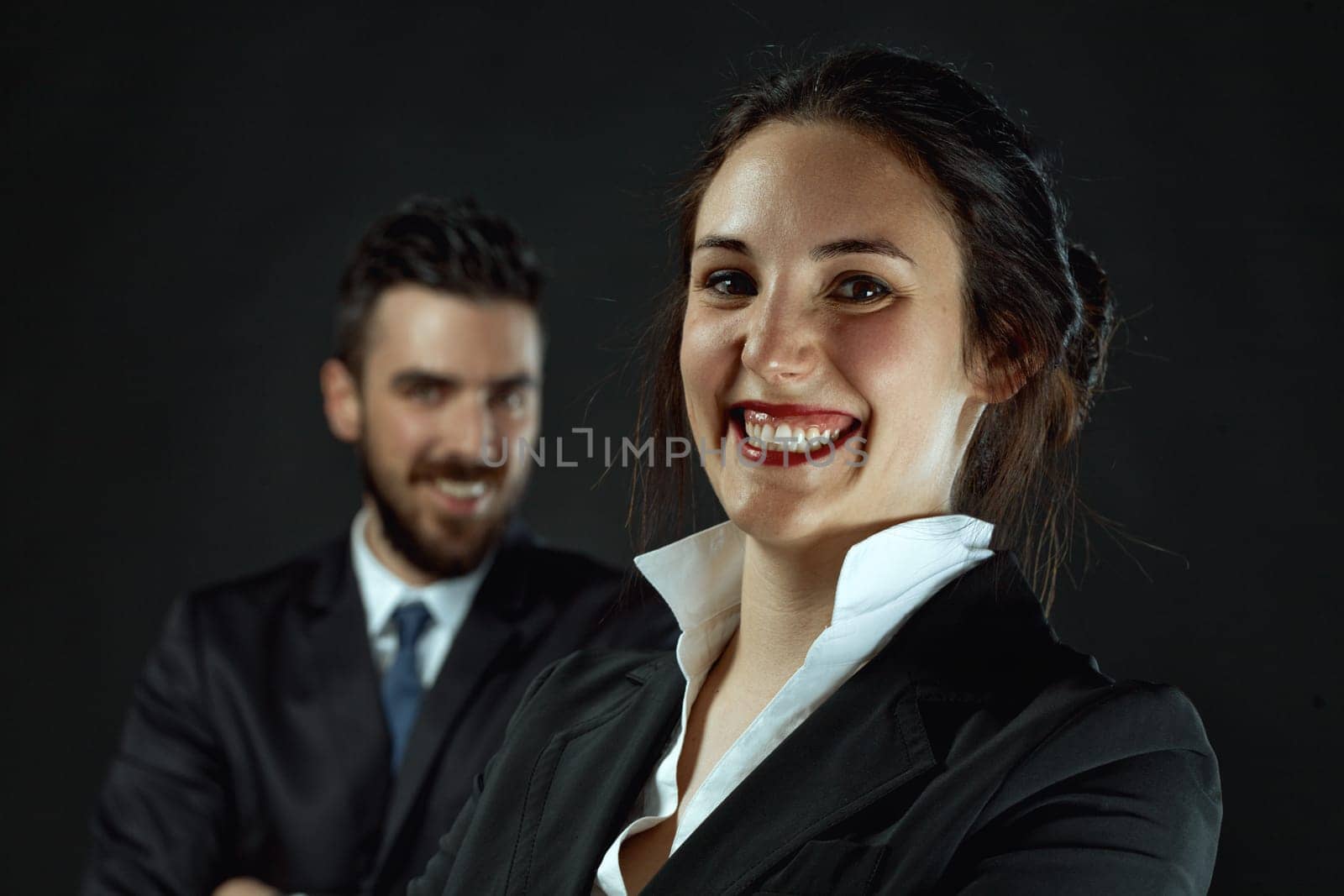 On a dark background, a sharp, prominent woman and a blurred, bearded man, both elegantly dressed in black business attire, epitomize professional assistance in various fields