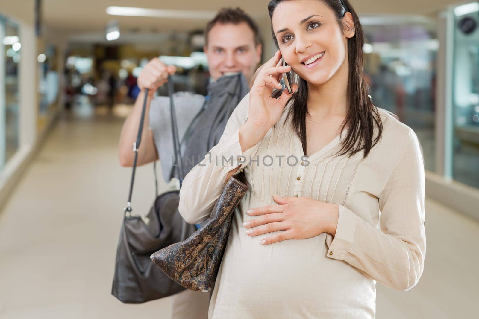 pregnant woman, engrossed in a phone call, is followed by her husband, burdened with her bags, symbolizing a dysfunctional relationship dynamic
